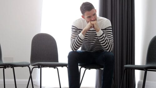 Patient seeking treatment for co-occurring mental and substance use disorders waits for a group therapy session to begin, seated with empty chairs on either side of him.