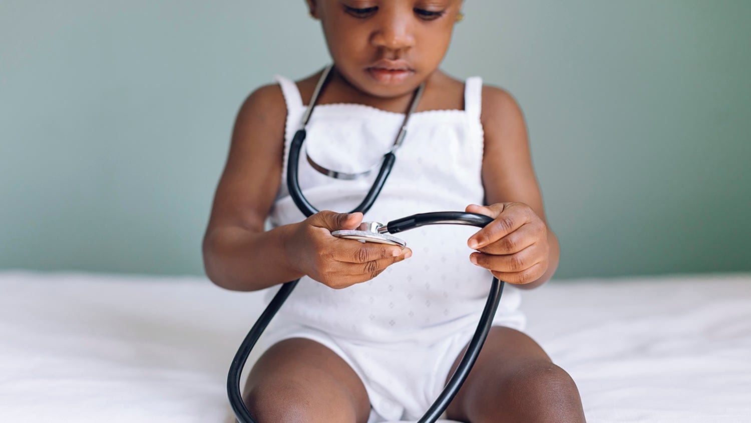 Young child sits on exam table looking at stethoscope in her hands