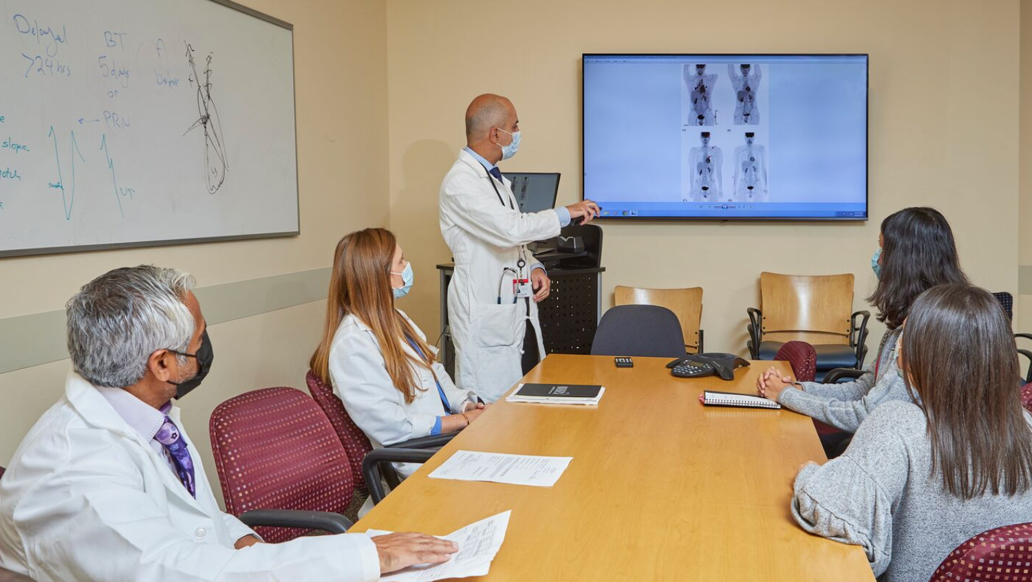 A group of people in white coats wearing masks are seated around a wooden conference table. A tall bald doctor is standing at the front of the room, clearly presenting patient scans. He and everyone at the table are masked.