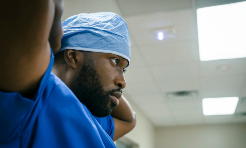 A black female doctor with long dark hair is wearing a white coat and holding a clipboard. A black man with short hair is seated on a patient bed and wearing a red striped polo shirt and jeans. They are both in a patient room.