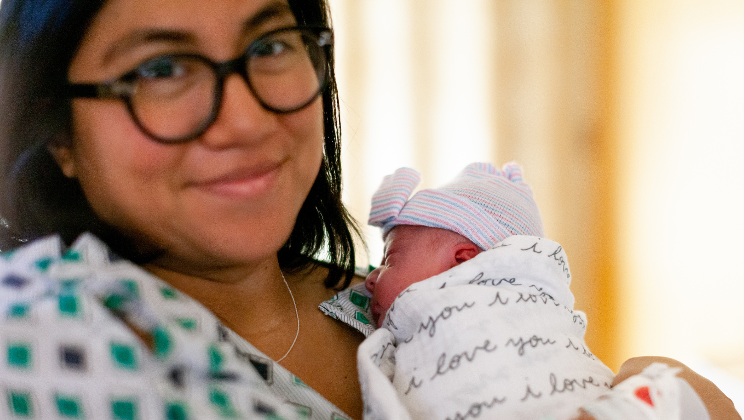 a doula with glasses smiles softly at the camera while holding a newborn to her chest