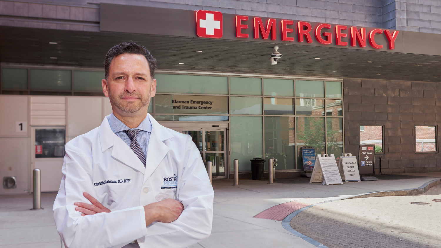 A male doctor with brown hair and brown eyes stands with his arms folded in front of the Emergency Department at Boston Medical Center. 