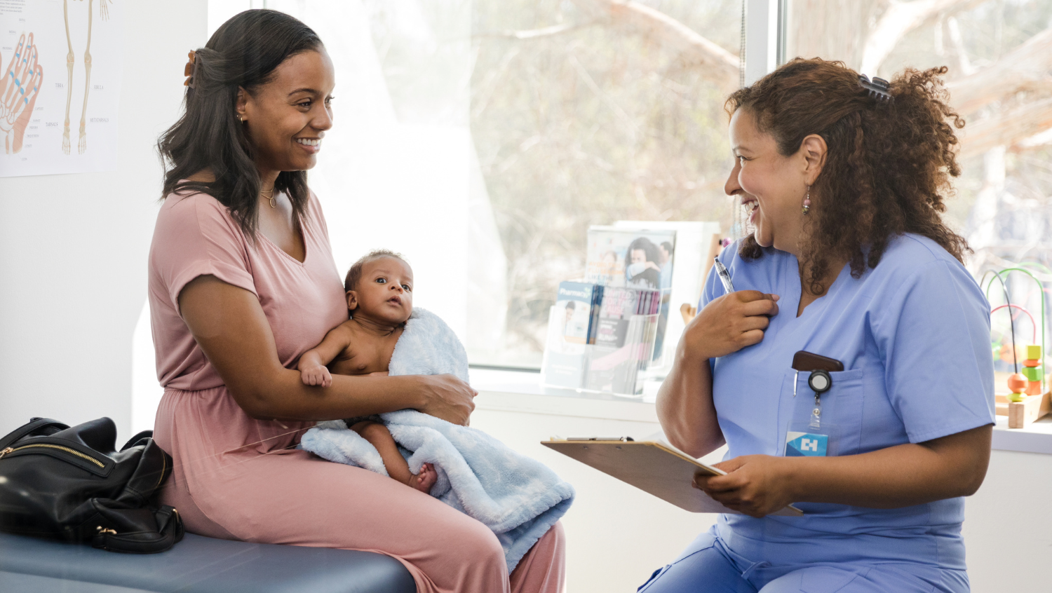 a black parent and her child are speaking with their healthcare provider. mother and provider are both smiling and looking at each other. the provider has thick, curly hair and tan skin and is wearing scrubs. the mother is wearing a pink jumpsuit