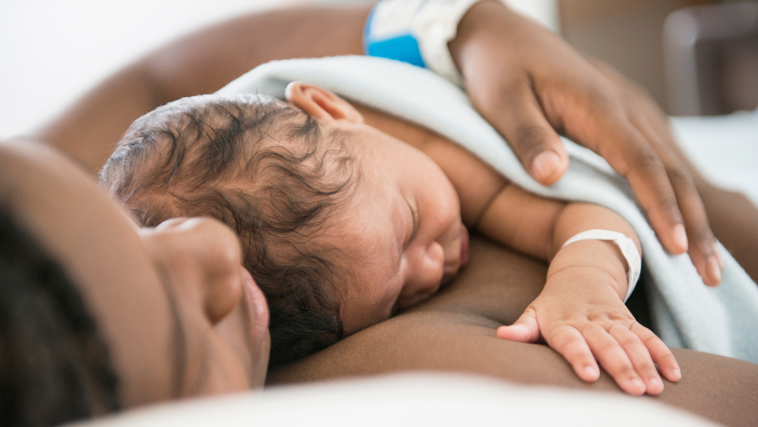 A Black woman sleeping with her newborn baby on a hospital bed. The baby's is lying on top of the mother and the mother is resting her hand gently over the baby's back. The baby has short, dark hair.  