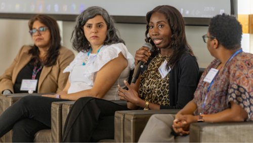 A Black woman with long brown hair is seated in a chair and speaking into a microphone. Two women sit to her right and one woman sits to her left. All of them are listening to her speak.