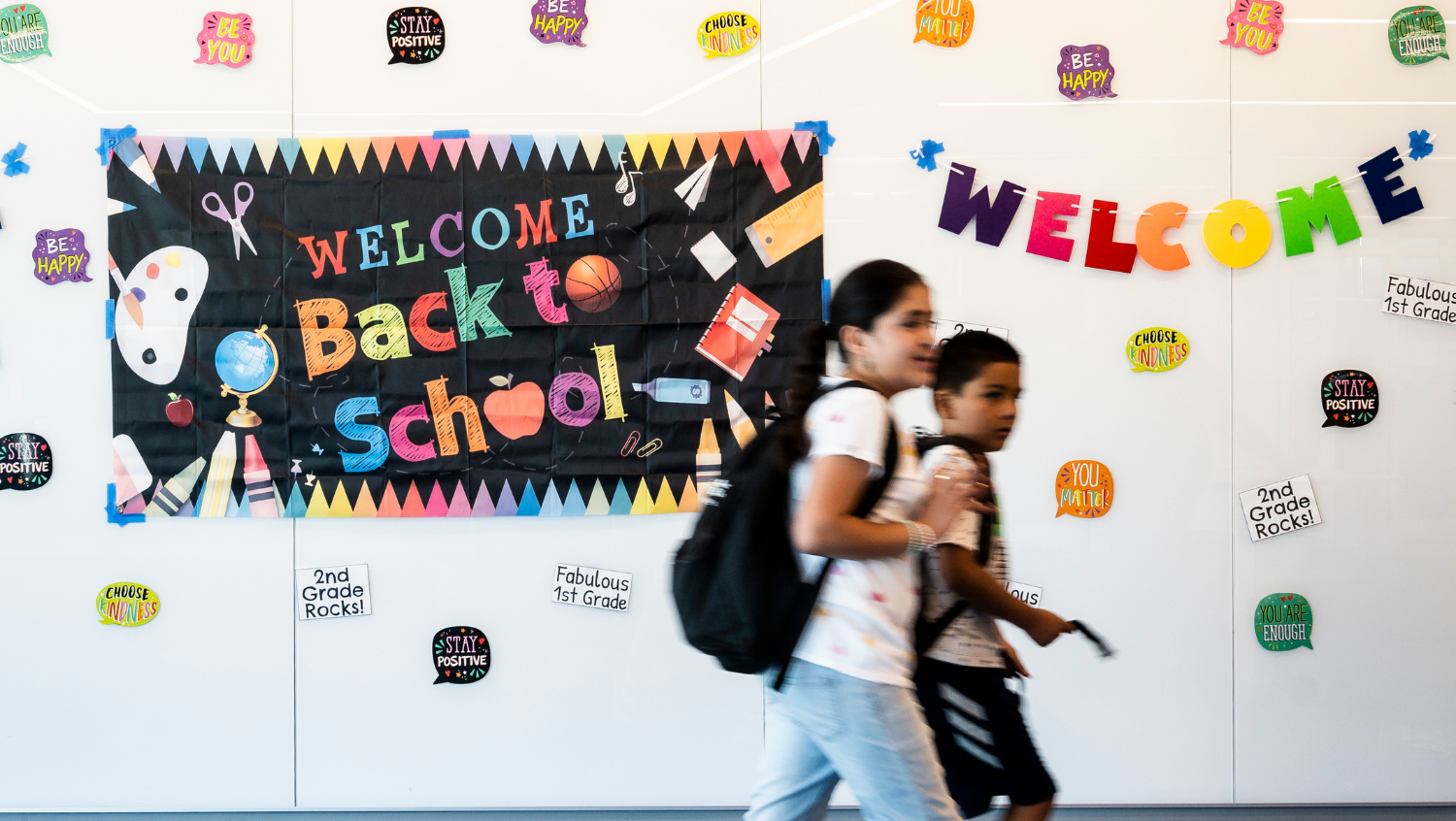 two students wearing backpacks walk down a school hallway decorated for the start of school. There are signs that say welcome back to school and posters with encouraging words on them.