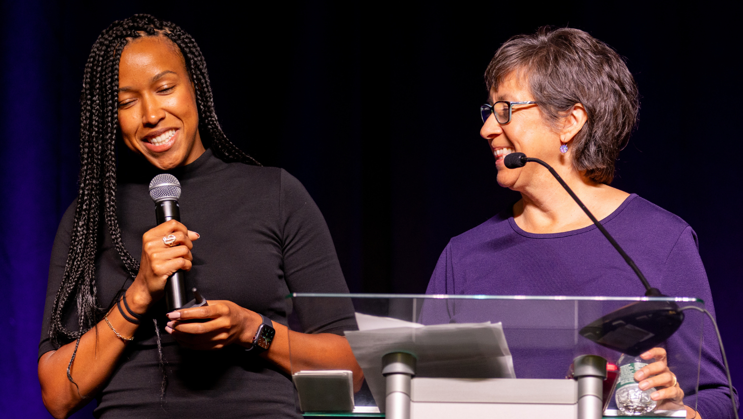 a black woman and a white woman each speak into the microphone, the black woman is looking down and smiling. the white woman is looking at the black woman and smiling