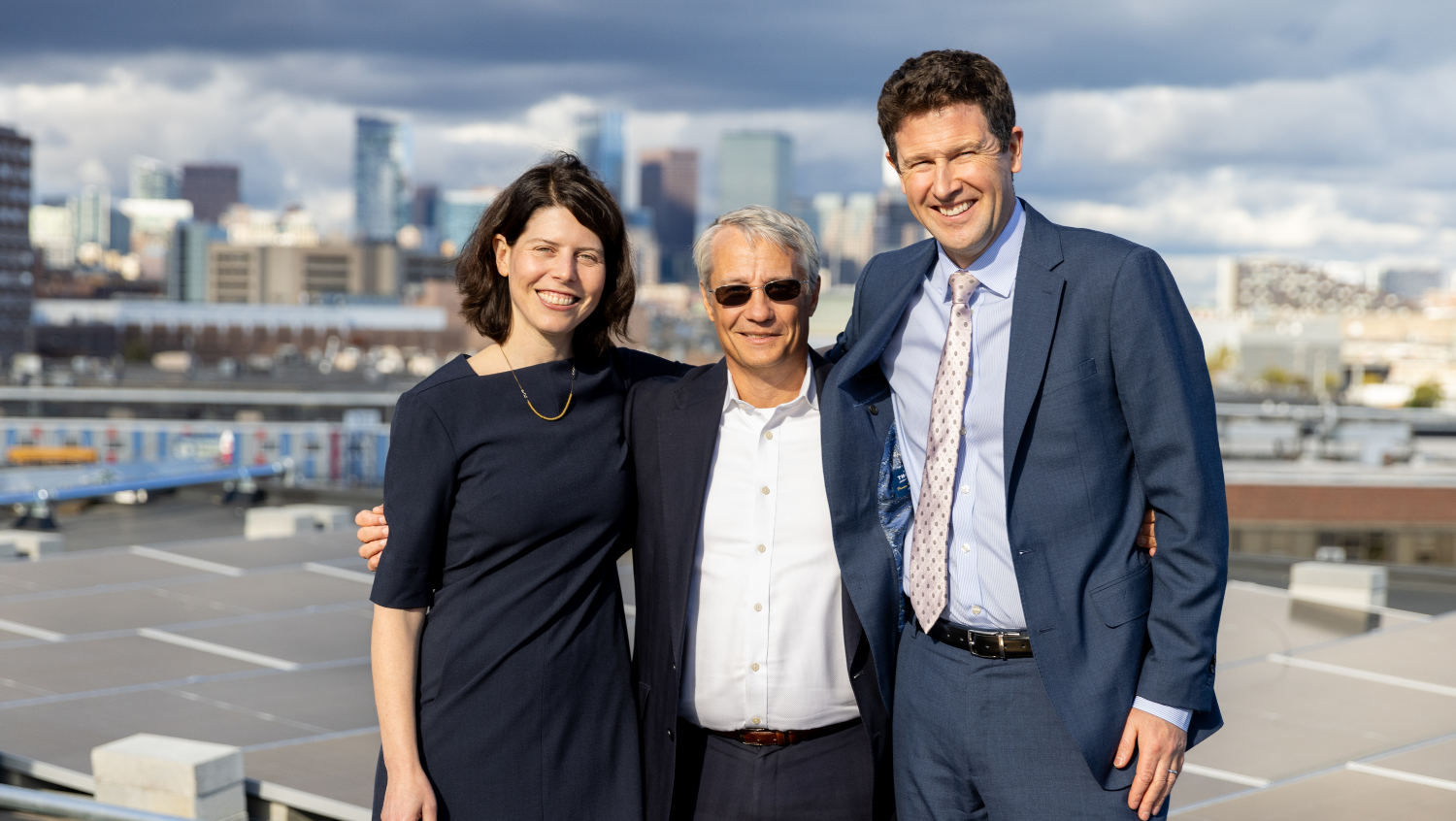 three white people: Anna Goldman, MD, MPA, MPH, Bob Biggio, Alastair Bell, MD, MBA are smiling and looking directly at the camera posing in front of a solar array with a faint view of the Boston skyline in the background. 
