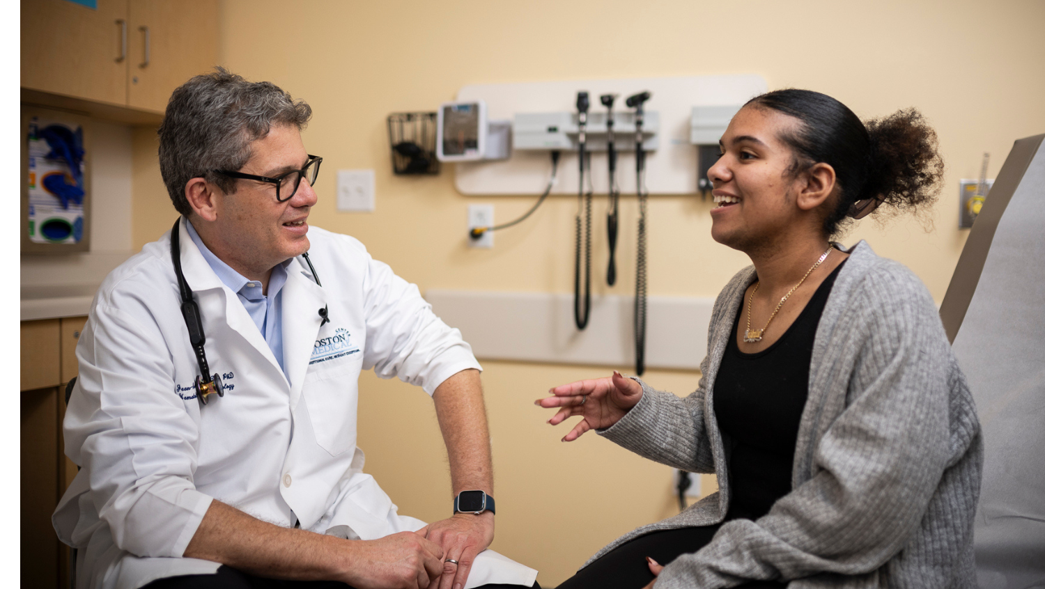 a doctor — a white man with glasses and a white lab coat — is sitting with his patient, a Black woman, who is smiling and talking with her hands. 