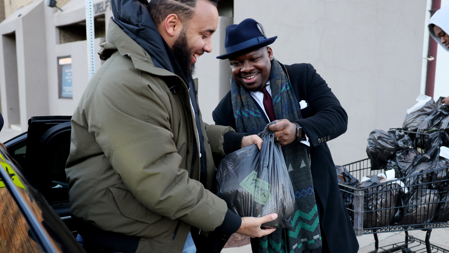 Two Black men are smiling at one another. The man facing the camera is wearing a top hat and carrying a frozen turkey in a bag as he hands it to the other man. 