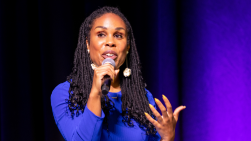 Uche Blackstock, a Black woman with locs is speaking into a microphone. She is wearing a blue dress and the backdrop is a royal purple.