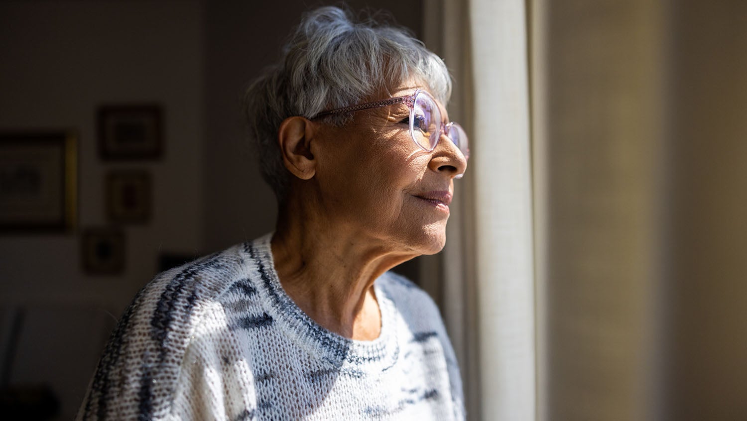 multiracial elderly woman wearing glasses and a sweater looking out a window