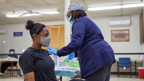 A Black nurse wearing a blue floral print scrubcap and a navy shirt vaccinates a young Black woman wearing a black t shirt. Both people are wearing masks. 