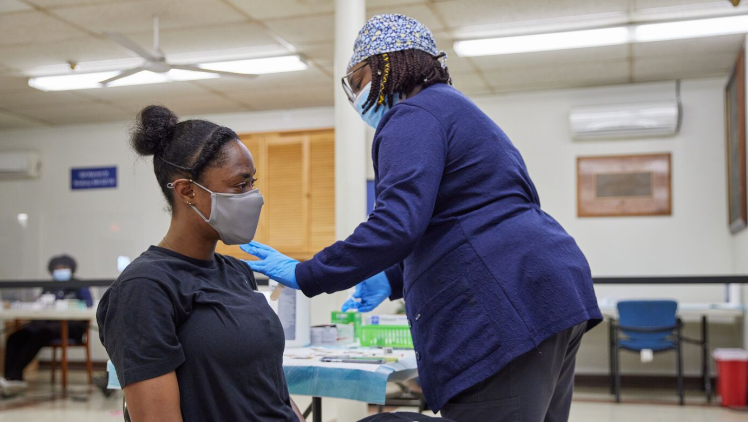A Black nurse wearing a blue floral print scrubcap and a navy shirt vaccinates a young Black woman wearing a black t shirt. Both people are wearing masks. 