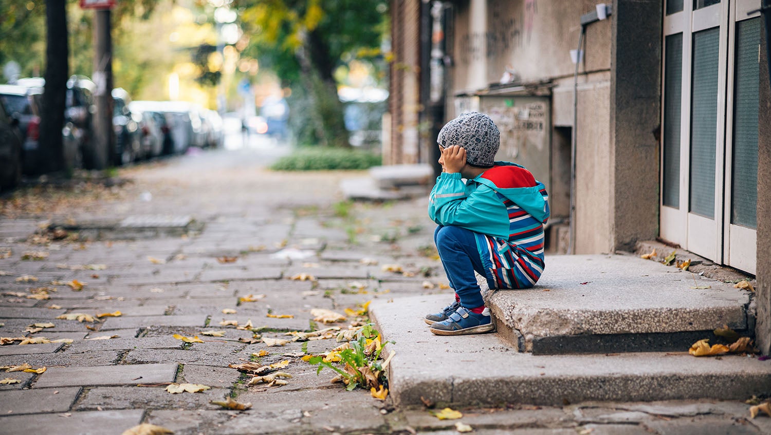 a little boy wearing a hat sits on a stoop outside a door on a city street, looking sad