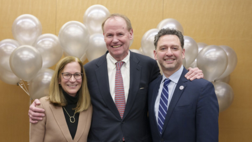 A group of three people smiling and facing the camera. On the right is a short white woman with caramel colored hair and glasses, next to her us a white man in a suit, next to him is a light skinned Latino man, also wearing a suit.