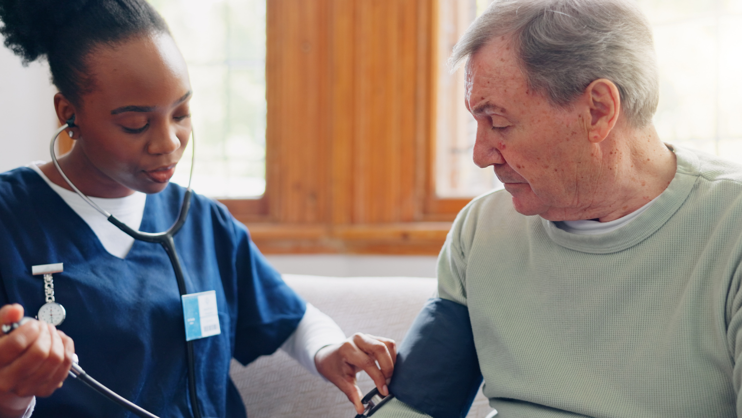 A black woman with scrubs and a stethoscope is taking the blood pressure of an older white man on a white couch in front of two open windows. He is clearly in his home.  