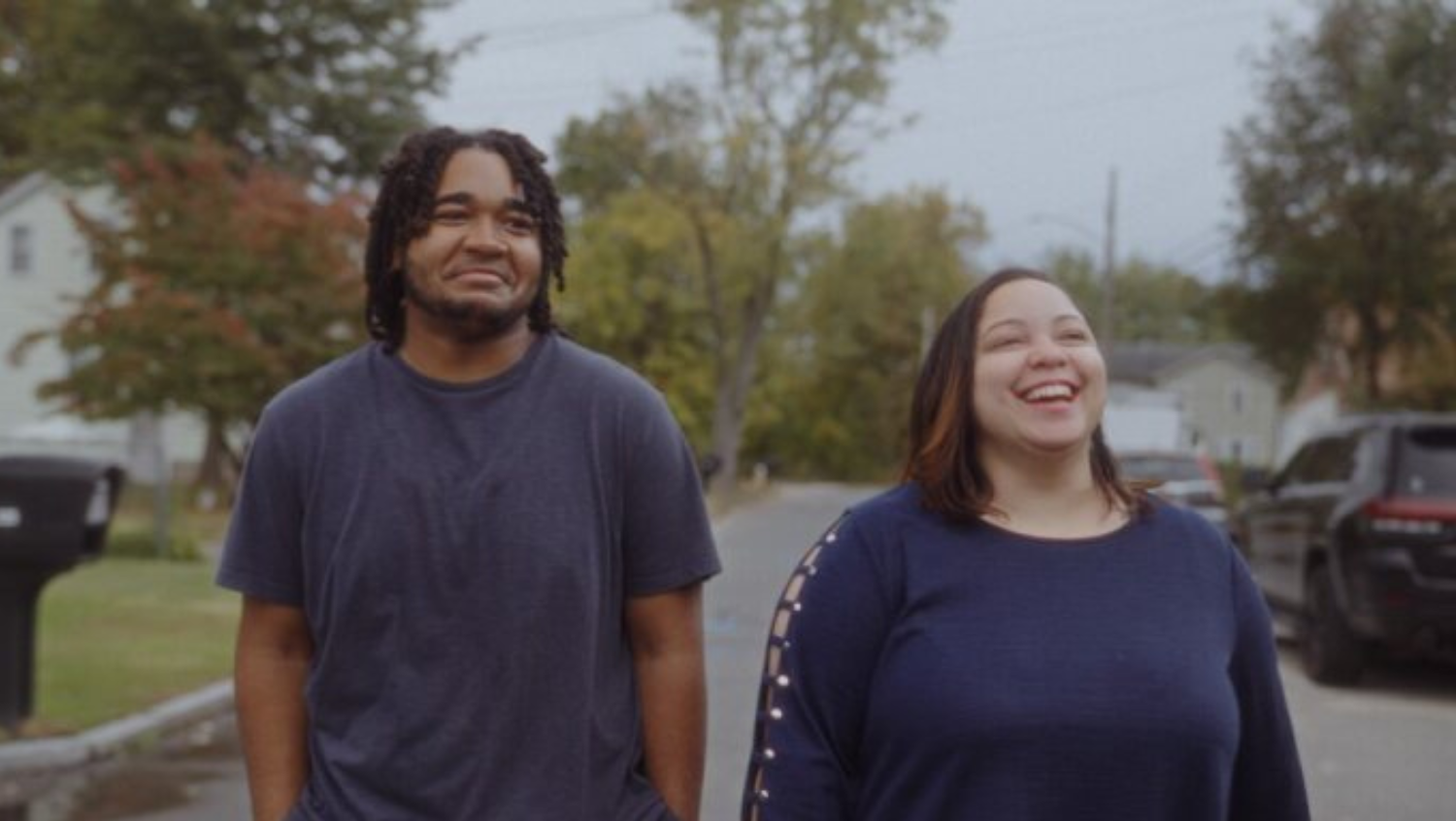 Two people: a young Black boy with dreadlocks and his mother, a light-skinned woman with straight hair are walking on a suburban street on a cloudy day. They are both laughing