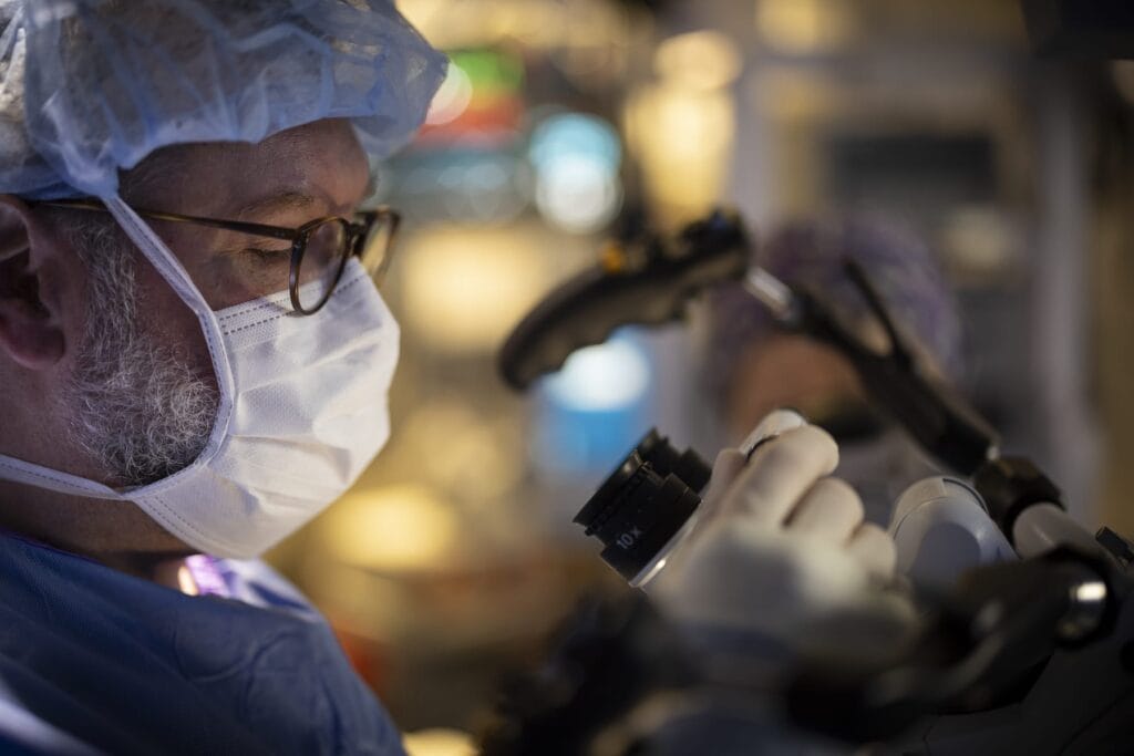 a doctor looking into a machine in the operating room for skull base surgery, wearing scrubs and a mask.