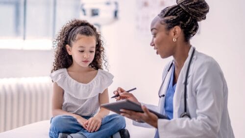 a young girl with curly hair sits on a bed in an exam room talking to a doctor holding a notepad.