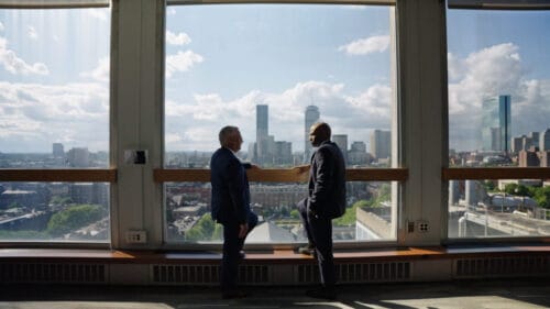 two men in suits standing in front of floor to ceiling windows overlooking boston skyline