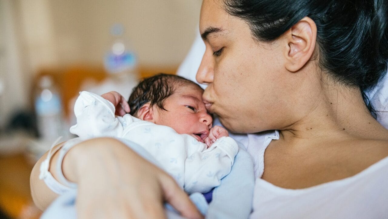 up close picture of a woman in a hospital bed kissing her newborn baby on the forehead