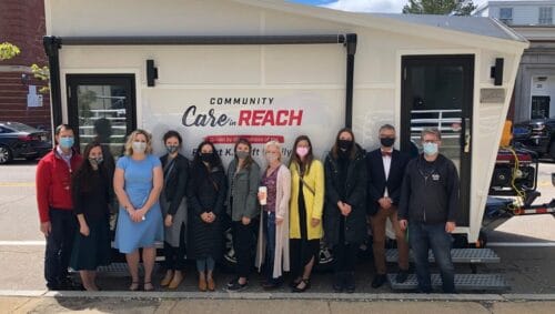 a group of varied people in medical masks standing in front of a mobile opioid use disorder treatment van that says community care in reach