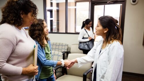 young girl with curly hair and her mother approaching a doctor in a white lab coat in a waiting room