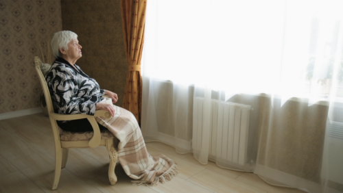 an elderly person in a hardback chair sitting with a blanket and looking out a large window