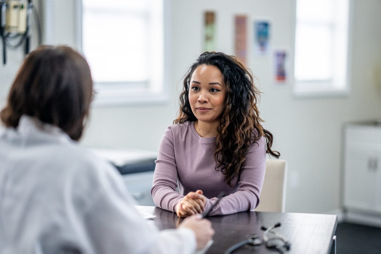 A young woman of mixed race, sits with her female doctor as they discuss her health concerns.  The patient is dressed casually and has a neutral expression on her face.