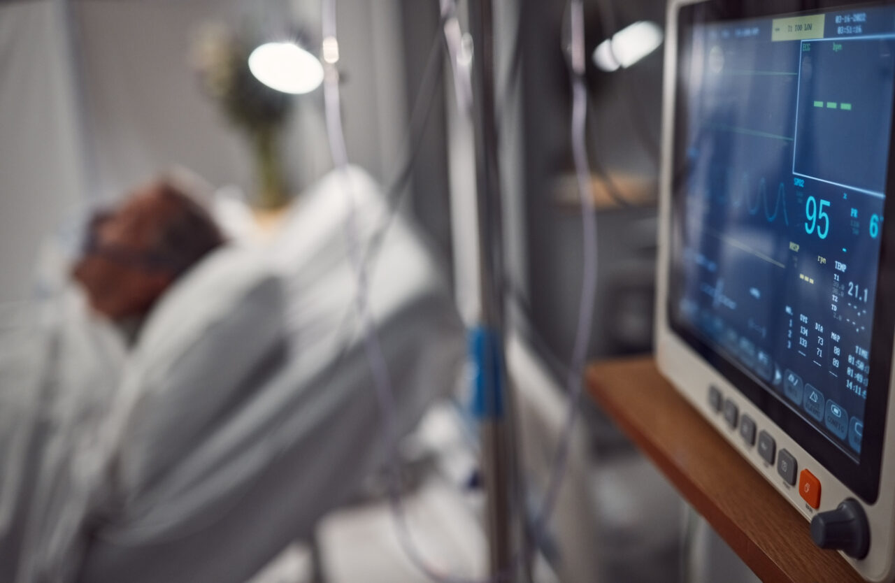 vital sign and pupillometry monitoring machines in the foreground with a patient blurred in an ICU bed in the back