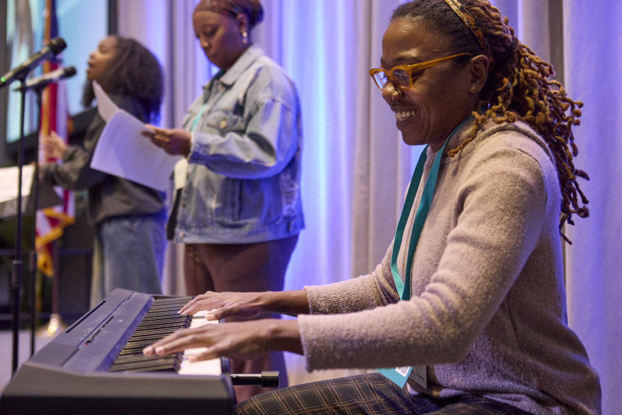 black woman sits smiling at a keyboard while perfomers sing on stage behind her
