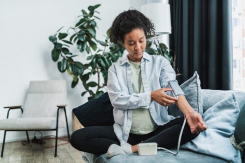young black woman sitting on her couch, pregnant using at home remote blood pressure monitor